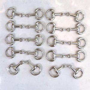 May include: A collection of silver-colored horse bits, each with a D-ring on either side and a central joint. The bits are arranged in rows and columns, with a few angled to show their flexibility. The background is a light, possibly white, surface.