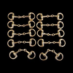 May include: An array of gold-colored horse bits, each with a D-ring design. The bits are arranged against a black background, showcasing their reflective, metallic finish. The image highlights equestrian equipment.