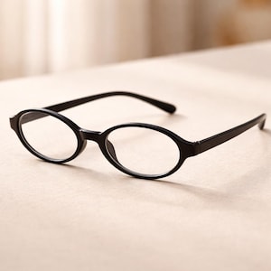 Puede incluir: Gafas ovaladas negras con lentes transparentes. Las monturas son de color negro sólido y las varillas son finas y rectas. Las gafas están sobre un fondo neutro, resaltando su diseño elegante.