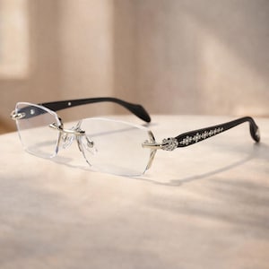 Puede incluir: Gafas sin montura con lentes transparentes y patillas negras. Las patillas están adornadas con un patrón de diamantes y detalles plateados decorativos. Las gafas tienen un diseño moderno y elegante, adecuado para el uso diario.