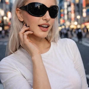 May include: Black oval sunglasses with a thick frame. The sunglasses reflect the surrounding environment. The person is wearing a white long-sleeve shirt. The person has blonde hair and a nose ring.