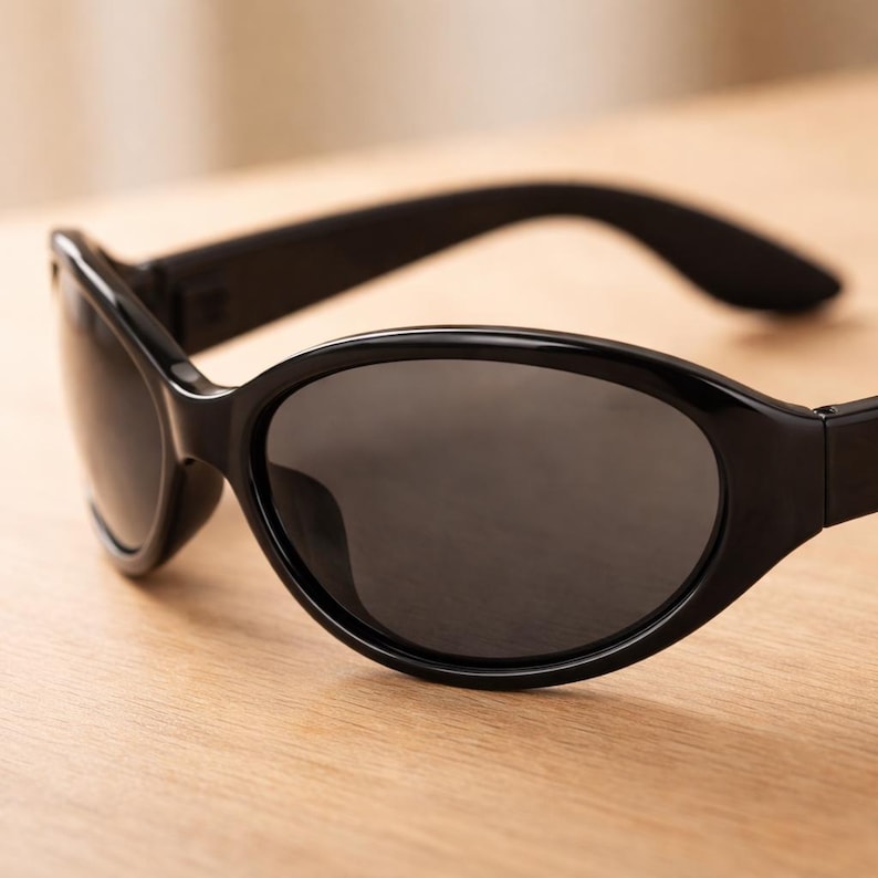 May include: Black sunglasses with oval lenses and a thick, glossy frame. The sunglasses are set against a light-colored wooden surface. The lenses are dark, providing shade and protection from the sun.