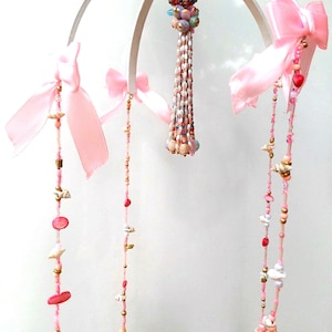 Puede incluir: Móvil hecho a mano con un marco blanco, lazos rosas y hebras de cuentas. Las hebras están decoradas con conchas marinas, cuentas rosas y detalles dorados. Una esfera de cuentas y una borla cuelgan del centro.