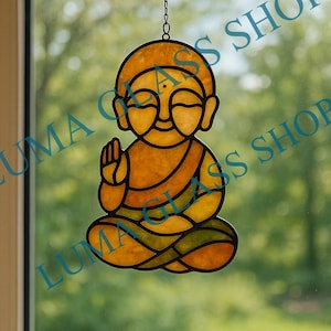 Pode incluir: Estatueta de Buda em vitral em pose sentada, com expressão serena e mão levantada. A obra apresenta tons de amarelo, laranja e verde, delineados em preto. A estatueta é suspensa por uma corrente, adequada para pendurar como peça decorativa.