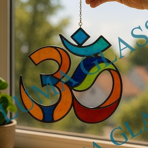 May include: A stained glass Om symbol in orange, blue, green, and red, hanging from a chain. The symbol is held up in front of a window. The text "LUMA GLASS" is visible in the lower right corner.