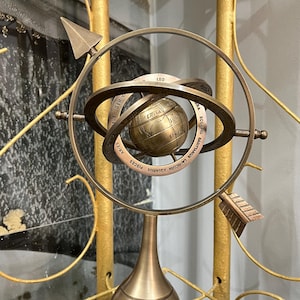 Puede incluir: Una esfera armilar de bronce con símbolos del zodiaco y un globo central. La esfera tiene elementos en forma de flecha y está montada sobre una base decorativa. El fondo presenta elementos metálicos dorados y una pared texturizada.