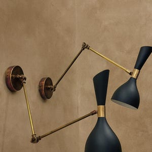 Puede incluir: Dos apliques de pared negros y dorados con brazos ajustables. Los apliques tienen un diseño vintage, con una pantalla azul oscuro y detalles dorados. Los soportes de pared son circulares y tienen un acabado bronce. Las lámparas están montadas en una pared marrón.
