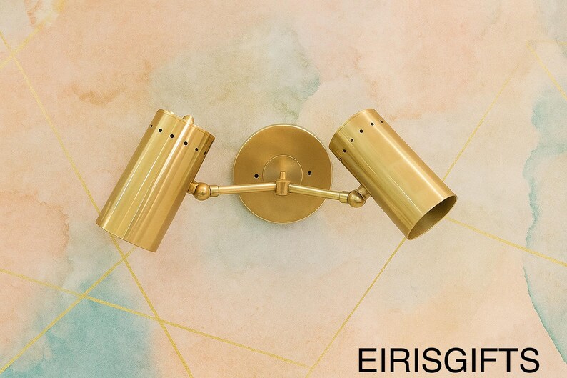 Op de afbeelding: Een messingkleurige wandlamp met twee lichten. De armatuur heeft twee cilindrische lichtkoppen met kleine perforaties, verbonden met een centrale cirkelvormige basis door verstelbare armen. De lamp is gemonteerd op een muur met een aquarelontwerp. De tekst "EIRISGIFTS" staat onderaan.