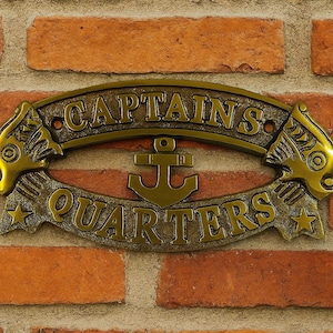 Pode incluir: Uma placa de metal bronzeada com as palavras "CAPTAINS QUARTERS", com uma âncora no centro e elementos decorativos. A placa está montada numa parede de tijolos vermelhos.