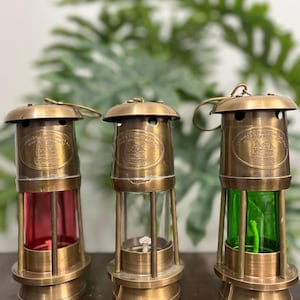 Op de afbeelding: Drie antieke messing mijnwerkerslampen met glazen cilinders. Eén lamp heeft een rode glazen cilinder, één is helder en de derde heeft een groene glazen cilinder. Elke lamp heeft een messing bovenkant met een handvat en een voet.