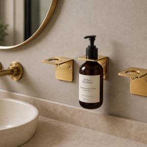 May include: Three brushed gold metal wall-mounted holders with L-shaped brackets. One holder supports a dark amber glass bottle with a black pump dispenser, labelled Atelier Savonnie Sandalwood & Vetiver Body Wash, 300ml 10.1 fl oz.