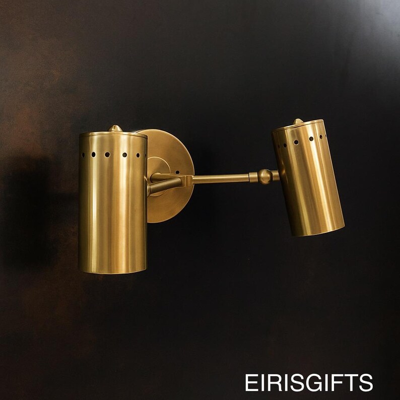 Op de afbeelding: Een messingkleurige wandlamp met twee cilindrische armaturen. Elk armatuur heeft een reeks kleine gaatjes aan de bovenkant. De lamp is gemonteerd op een cirkelvormige basis en heeft een verbindingsarm. De tekst "EIRISGIFTS" staat onderaan.