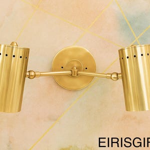Op de afbeelding: Een messing wandlamp met twee verstelbare cilindrische lichtkoppen. De lamp heeft een cirkelvormige basis en is bevestigd aan een muur met een aquarel-achtige achtergrond. De tekst "EIRISGIFTS" staat onderaan.