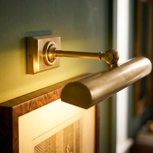 May include: A wall-mounted picture light with a brushed brass finish, featuring a rectangular base plate, a horizontal adjustable arm, and a cylindrical light housing that casts a warm glow downward onto a framed artwork below.