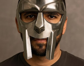 May include: A silver-colored metal mask with a distinctive design, featuring a prominent nose and vertical bars extending from the mouth area. The mask covers the upper face, with visible eyes. The person is wearing a dark gray t-shirt.