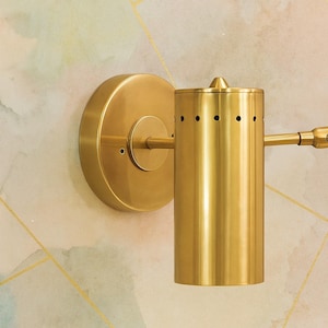 Op de afbeelding: Een goudkleurige wandlamp met een cilindrische lampenkap en een cirkelvormige basis. De lamp heeft kleine gaatjes aan de bovenkant en een kleine arm die naar rechts uitsteekt. De achtergrond is een patroonmuur met gouden lijnen en de tekst "EIRISGIFTS".