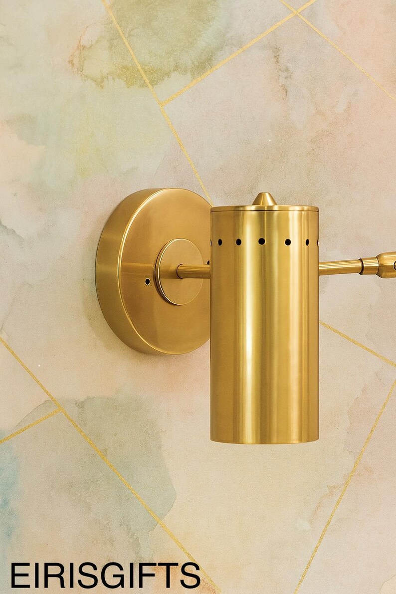 Op de afbeelding: Een goudkleurige wandlamp met een cilindrische lampenkap en een cirkelvormige basis. De lamp heeft kleine gaatjes aan de bovenkant en een kleine arm die naar rechts uitsteekt. De achtergrond is een patroonmuur met gouden lijnen en de tekst "EIRISGIFTS".
