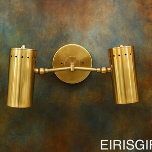 Op de afbeelding: Een messing wandlamp met twee cilindrische armaturen. Elk armatuur heeft een reeks kleine gaatjes aan de bovenkant. De lamp is gemonteerd op een ronde achterplaat en verbonden door een horizontale arm. De achtergrond is een gevlekt mengsel van bruin, blauw en groen.