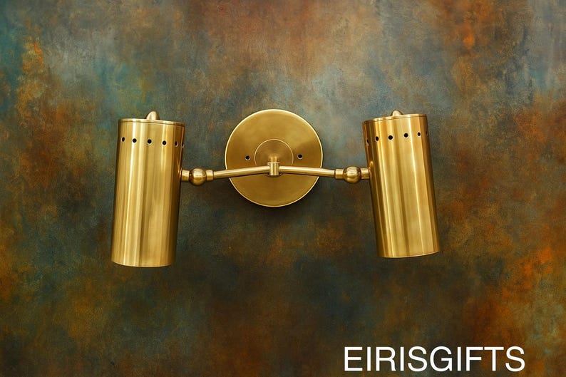 Op de afbeelding: Een messing wandlamp met twee cilindrische armaturen. Elk armatuur heeft een reeks kleine gaatjes aan de bovenkant. De lamp is gemonteerd op een ronde achterplaat en verbonden door een horizontale arm. De achtergrond is een gevlekt mengsel van bruin, blauw en groen.