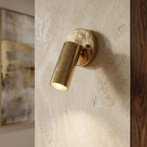 May include: A brass-colored wall-mounted spotlight fixture. The cylindrical light is angled downward, emitting a warm glow. The fixture is attached to a circular base, mounted on a textured, light-colored wall. A dark wood panel is visible on the right.
