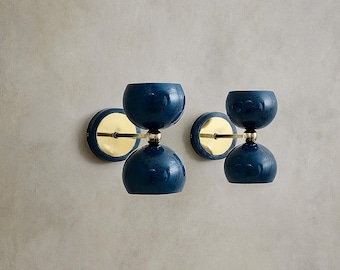 May include: Two wall sconces with a dark blue, hourglass-shaped design. Each sconce features a gold-colored circular backplate and a matching gold rod connecting the two blue bulbous sections. The sconces are mounted on a light gray wall.