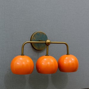May include: A modern wall sconce with three orange globe-shaped lights. The fixture has a brass-colored frame and a circular mounting plate. The sconce is mounted on a gray textured wall.
