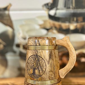 Puede incluir: Jarra de cerveza de madera con un diseño de árbol tallado. La jarra presenta un acabado de veta de madera natural, acentuado con bandas doradas. El asa es de madera clara. El diseño del árbol está grabado en la madera.