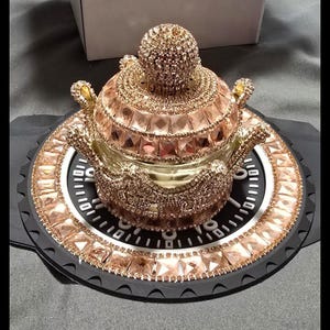 Könnte beinhalten: Ein dekorativer, roségoldfarbener Behälter mit Deckel, verziert mit glitzernden Kristallen. Der Behälter steht auf einer schwarz-weißen, kreisförmigen Basis, die ebenfalls mit Kristallen verziert ist. Das Design ist aufwendig und luxuriös.