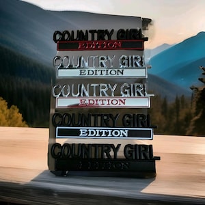 May include: A display of six "Country Girl Edition" badges in black, silver, and red. The text is in a clear, bold font. The background shows a mountain landscape with a scenic view.