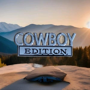 Puede incluir: Un emblema cromado "COWBOY EDITION" se muestra sobre un fondo de montañas y un atardecer. Debajo, un objeto marrón con detalles azules, posiblemente una funda de coche. El tema general sugiere el Oeste o la aventura al aire libre.