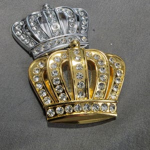 May include: Two decorative crowns, one silver and one gold, both adorned with clear rhinestones. The crowns feature arched supports and a rounded base, suggesting a regal theme. The gold crown is positioned beneath the silver one.