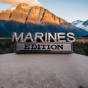 May include: A chrome emblem displaying the word "MARINES" above "EDITION". The emblem is set against a backdrop of a mountain range under a blue sky. The mountains are lit by the sun, creating a scenic view.