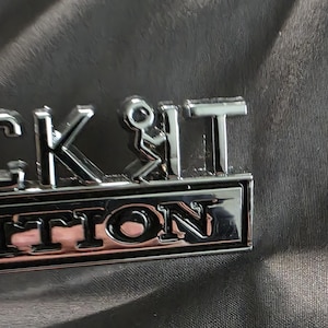 Könnte beinhalten: Ein verchromtes Metallabzeichen mit den Worten "FUCK IT EDITION" in Großbuchstaben. Der Teil "FUCK IT" zeigt eine Strichmännchen-Figur in der Mitte. Das Abzeichen ist vor einem dunklen, strukturierten Hintergrund platziert.