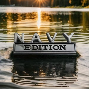 May include: A chrome emblem with the word "NAVY" above "EDITION" floats on water. The letters are raised and metallic, reflecting the sunlight. The background is a blurred view of water and a bright sunset.