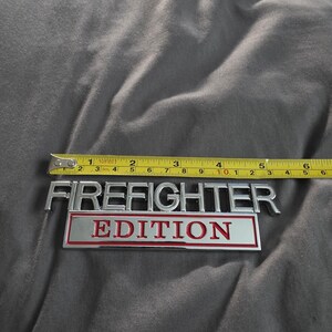 以下が含まれることがあります： 約12.7cmのクロムと赤色の「FIREFIGHTER EDITION」エンブレムが表示されています。エンブレムは黄色の巻尺で測定されています。テキストは太字のサンセリフ体です。