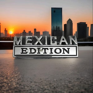 May include: A shiny chrome emblem displaying the text "MEXICAN EDITION" in capital letters. The emblem is set against a backdrop of a city skyline at sunset. The text is in a classic, readable font.