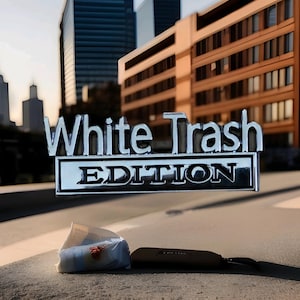 May include: A shiny chrome emblem displaying the text "White Trash EDITION" in a prominent font. The emblem is set against a blurred urban backdrop of buildings and a pavement. A small bag and a case are in the foreground.