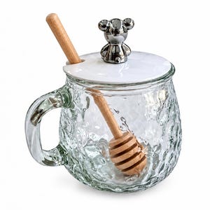 Op de afbeelding: Een doorzichtige glazen mok met een getextureerd, bobbelig oppervlak, voorzien van een wit deksel met daarop een zilverkleurig berenfiguurtje. Een houten honingdipper rust in de mok en steekt door een inkeping in het deksel naar buiten.