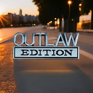 May include: Chrome "OUTLAW EDITION" emblem. The emblem features raised chrome lettering and a rectangular base with the word "EDITION" in black. The background shows a blurred street scene with a sunset.