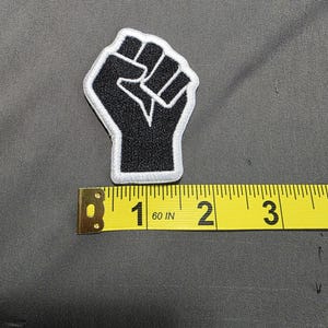 May include: Black embroidered patch featuring a raised fist with a white border. The patch is approximately 2 inches in height. The patch is on a gray fabric surface.
