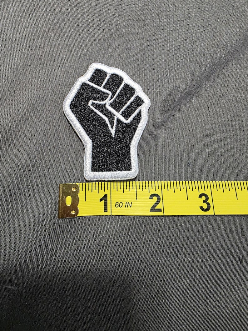 May include: Black embroidered patch featuring a raised fist with a white border. The patch is approximately 2 inches in height. The patch is on a gray fabric surface.