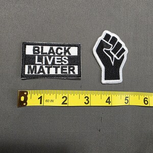 May include: Two embroidered patches: one rectangular with "BLACK LIVES MATTER" in white on black, and a black fist with a white outline. A yellow measuring tape shows the patches are approximately 2 inches and 1.5 inches in size.