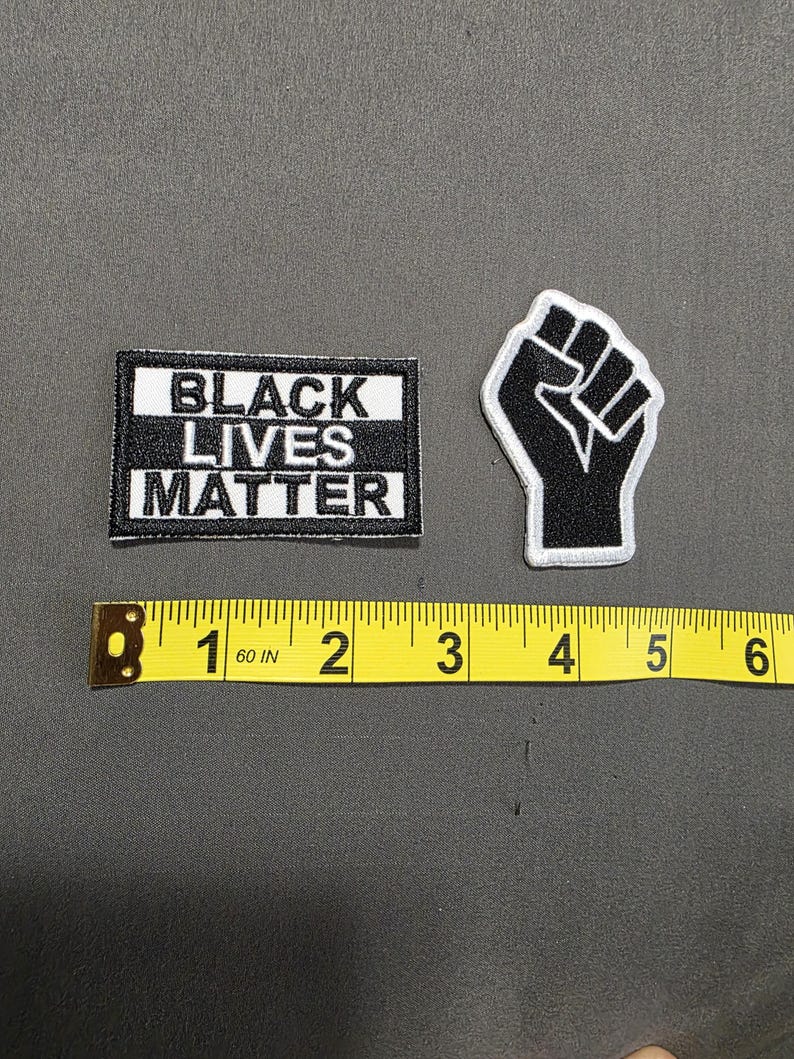May include: Two embroidered patches: one rectangular with "BLACK LIVES MATTER" in white on black, and a black fist with a white outline. A yellow measuring tape shows the patches are approximately 2 inches and 1.5 inches in size.