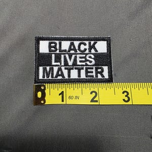 May include: A rectangular black and white patch with the text "BLACK LIVES MATTER". The patch is approximately 2 inches (5 cm) wide and is placed on a gray fabric surface. A yellow measuring tape is next to the patch.