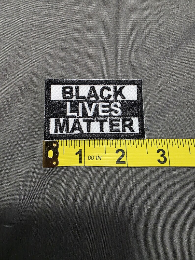 May include: A rectangular black and white patch with the text "BLACK LIVES MATTER". The patch is approximately 2 inches (5 cm) wide and is placed on a gray fabric surface. A yellow measuring tape is next to the patch.