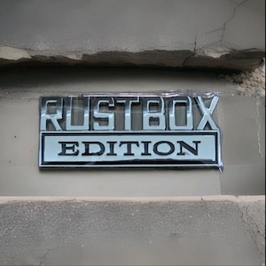 May include: A silver and black "RUSTBOX EDITION" emblem is affixed to a concrete surface. The emblem is rectangular with the words "RUSTBOX" in large, raised letters above the word "EDITION". The background is a light gray concrete.