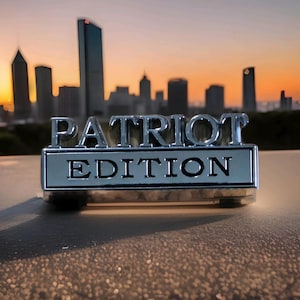 May include: A chrome emblem displaying "PATRIOT EDITION" set against a cityscape at sunset. The rectangular emblem has raised lettering, reflecting the warm colours of the sky and the silhouettes of buildings in the background.