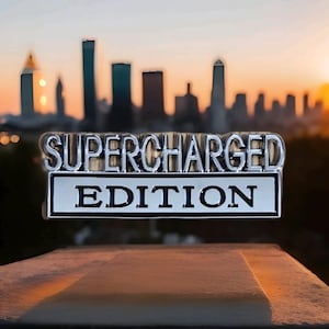 Peut inclure: Un emblème "SUPERCHARGED EDITION" chromé avec un fond blanc, sur fond de paysage urbain flou au coucher du soleil. L'emblème est monté sur une surface en béton, avec la silhouette de la ville en arrière-plan.