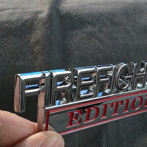 以下が含まれることがあります： 「FIRE FIGHTER」の文字が刻まれたクロム仕上げのエンブレム。その下に「EDITION」の文字が赤い文字で黒い背景に表示され、赤い縁で囲まれています。エンブレムは、暗く質感のある表面に対して保持されています。