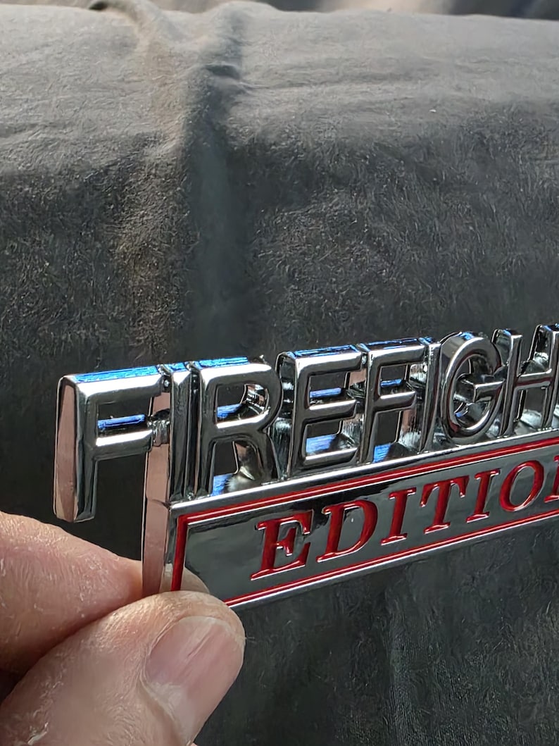 以下が含まれることがあります： 「FIRE FIGHTER」の文字が刻まれたクロム仕上げのエンブレム。その下に「EDITION」の文字が赤い文字で黒い背景に表示され、赤い縁で囲まれています。エンブレムは、暗く質感のある表面に対して保持されています。